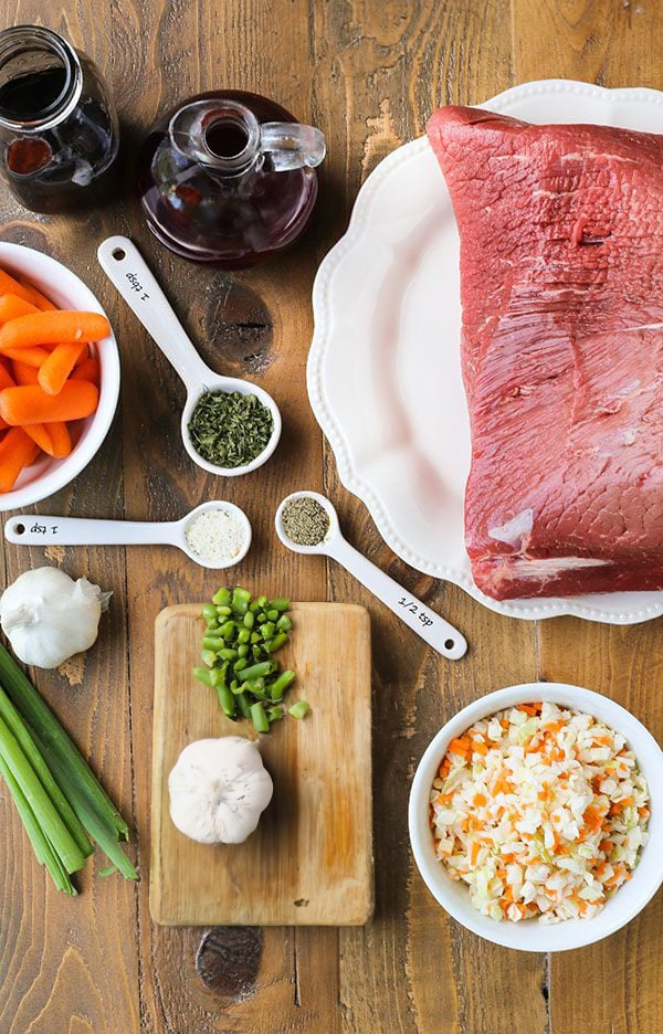 Top view of ingredients for Barbecue Honey Pot Roast, including a beef roast, baby carrots, green onions, garlic, and various seasonings, laid out on a wooden table.