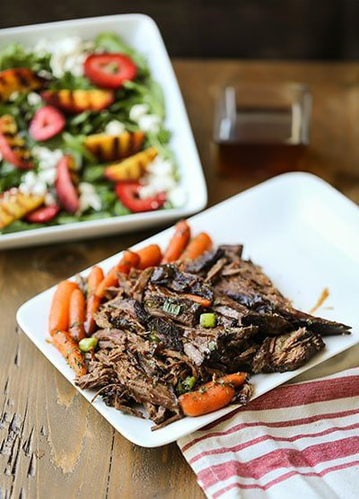 A plate of Barbecue Honey Pot Roast with carrots served next to a vibrant grilled peach and strawberry salad, displayed on a wooden table with a red-striped napkin.