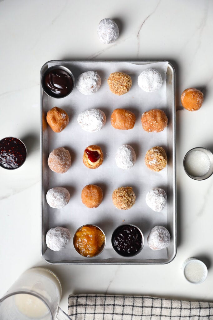 Freshly Made Donut Holes – Powdered, Glazed, and Coated Donut holes arranged on a tray with dipping sauces, including chocolate and fruit jam, placed on a marble countertop.