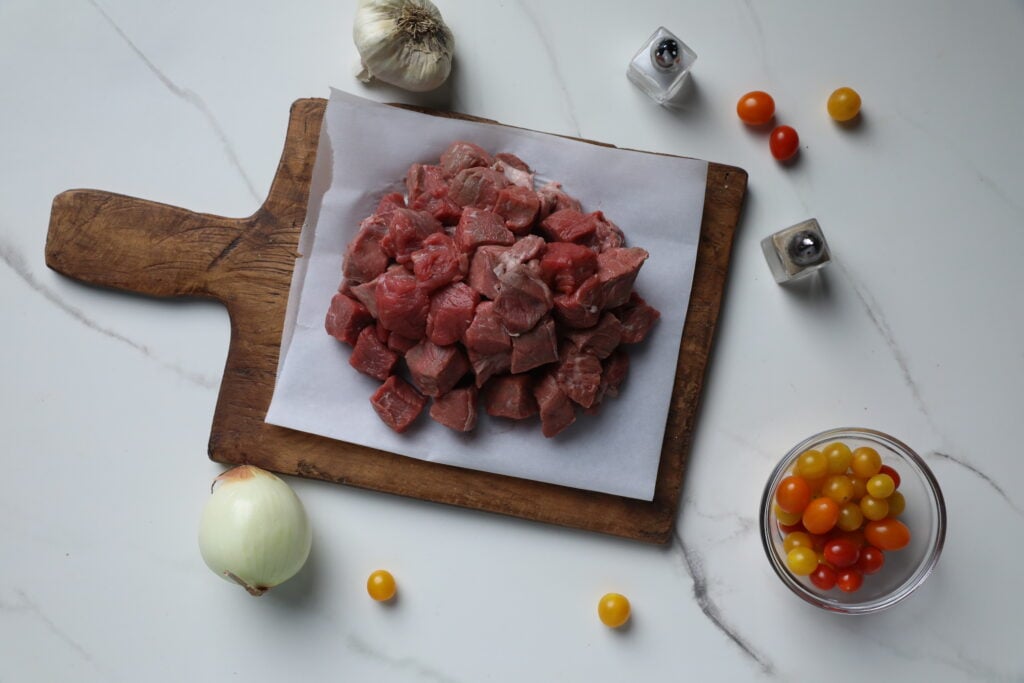 Tender Steak Bites on Parchment Paper with Fresh Ingredients Steak bites laid out for preparation with fresh ingredients like tomatoes, onion, and garlic on a white marble countertop.