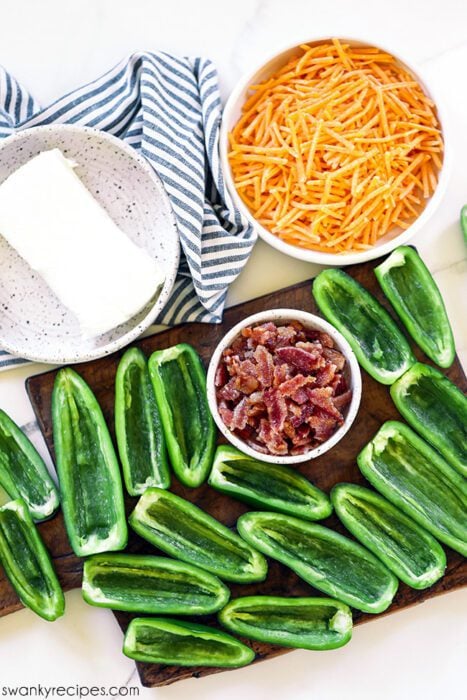 Jalapeno popper ingredients on a tabletop. Cheddar cheese, cream cheese, and bacon all in bowls with sliced jalapenos on the side.