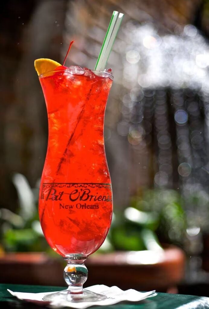 20 Must-Try Foods and Activities in New Orleans 17 The iconic Pat O'Brien's Hurricane glass, filled with the vibrant red Hurricane cocktail, stands on a table reflecting its rich, colorful history. The glass is tall, curved, and embossed with the Pat O'Brien's logo, showcasing the drink's strong association with New Orleans' festive bar culture.