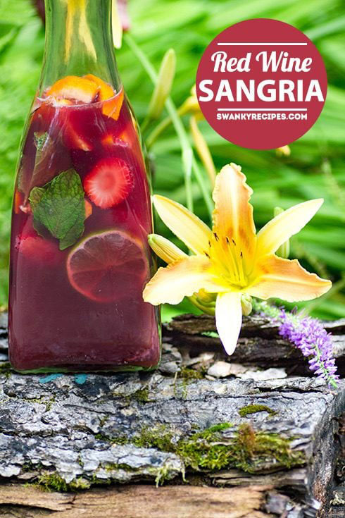 A tall glass bottle filled with vibrant red wine sangria, garnished with fresh fruit slices including oranges, strawberries, and a sprig of mint, sits on a rustic wooden surface. A bright yellow lily flower blooms beside the bottle, with lush green foliage in the background.