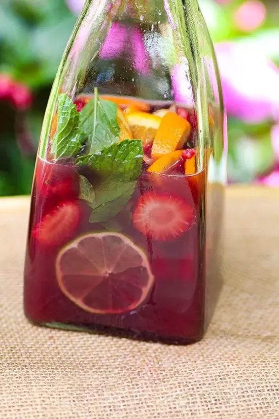 A close-up of a glass bottle filled with red wine sangria, showcasing slices of lemons, strawberries, and fresh mint leaves. The bottle is set against a background of bright green foliage and pink flowers, highlighting the refreshing and colorful nature of the drink.
