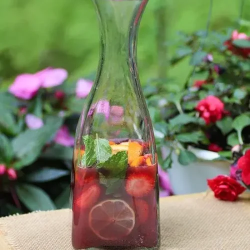 A tall glass bottle filled with red wine sangria, featuring slices of lemon, strawberries, and fresh mint leaves. The bottle is placed on a burlap-covered surface, with a background of lush green foliage and pink flowers, highlighting the refreshing and colorful nature of the drink.