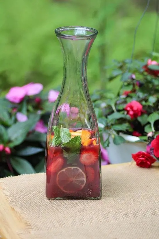 A tall glass bottle filled with red wine sangria, featuring slices of lemon, strawberries, and fresh mint leaves. The bottle is placed on a burlap-covered surface, with a background of lush green foliage and pink flowers, highlighting the refreshing and colorful nature of the drink.