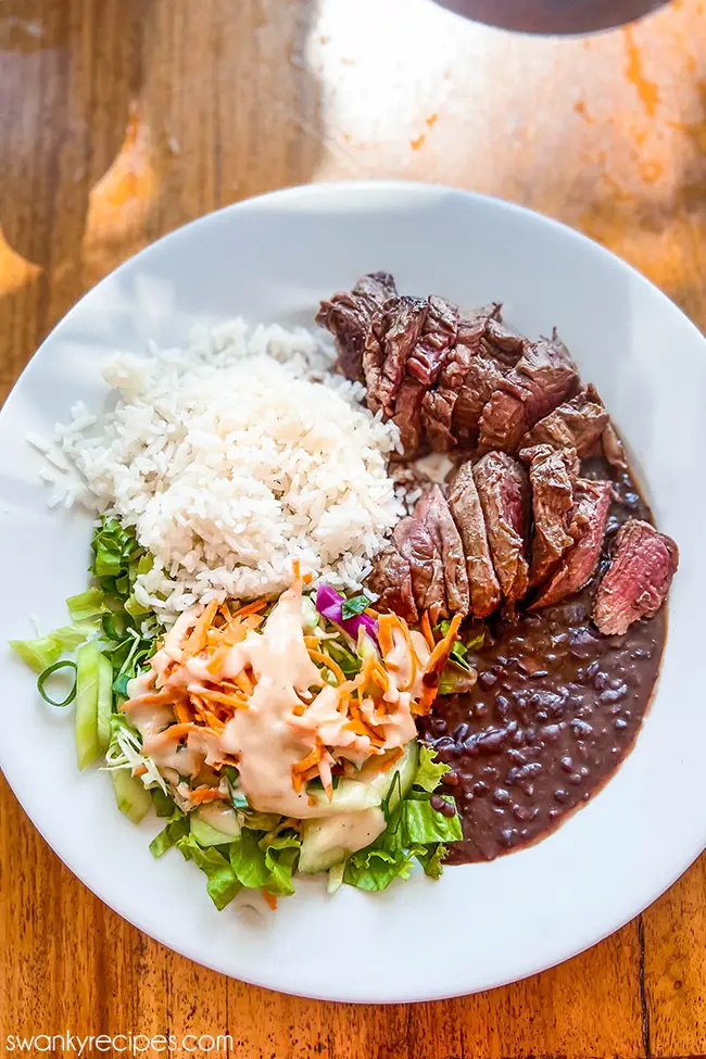 A plate of Costa Rican food with grilled steak, white rice, black beans, and a fresh salad with creamy dressing.