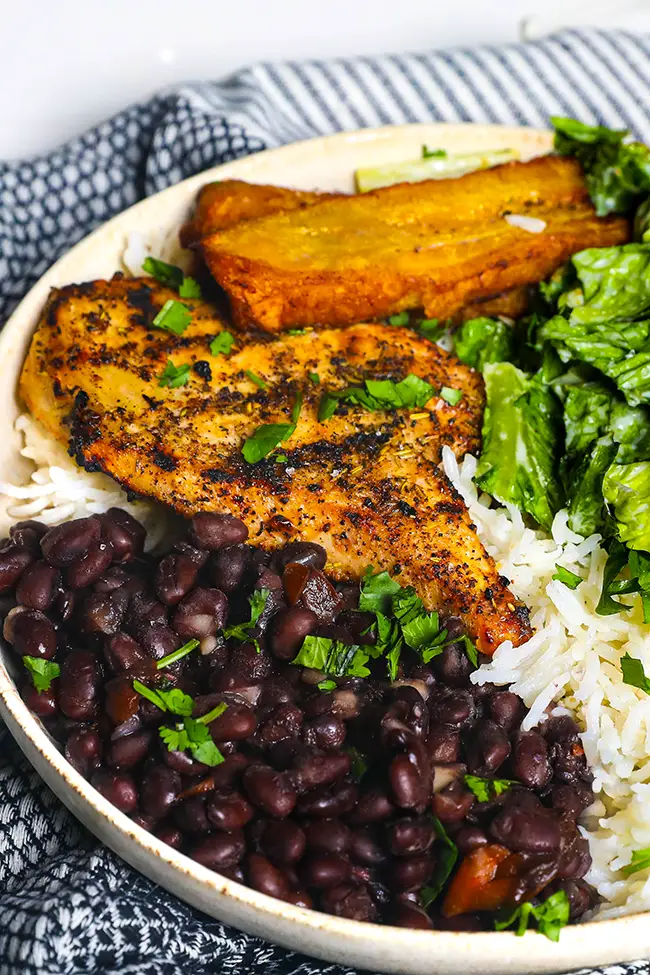 A plate of Costa Rican black beans served with grilled chicken, fried plantains, white rice, and fresh greens.